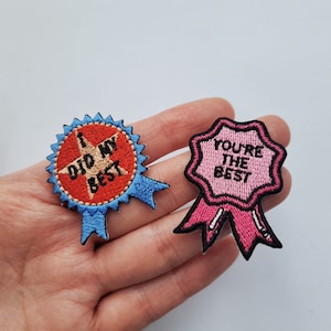 Peut inclure: Deux écussons brodés. L'un est un cercle rouge avec une étoile et le texte "I DID MY BEST" sur un ruban bleu. L'autre est une forme rose festonnée avec le texte "YOU'RE THE BEST" sur un ruban rose.