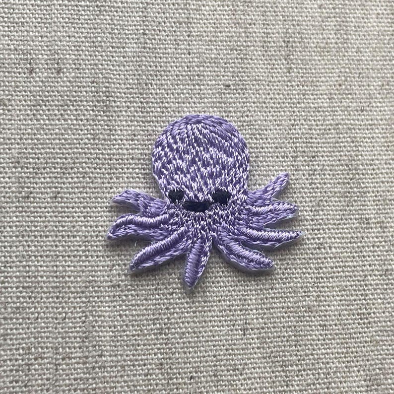 Pu&ograve; includere: Una piccola toppa ricamata a forma di polpo nei toni del lavanda e del bianco. Il polpo ha un volto sorridente e otto tentacoli dettagliati. La toppa &egrave; probabilmente progettata per essere cucita su tessuto, aggiungendo un elemento decorativo.