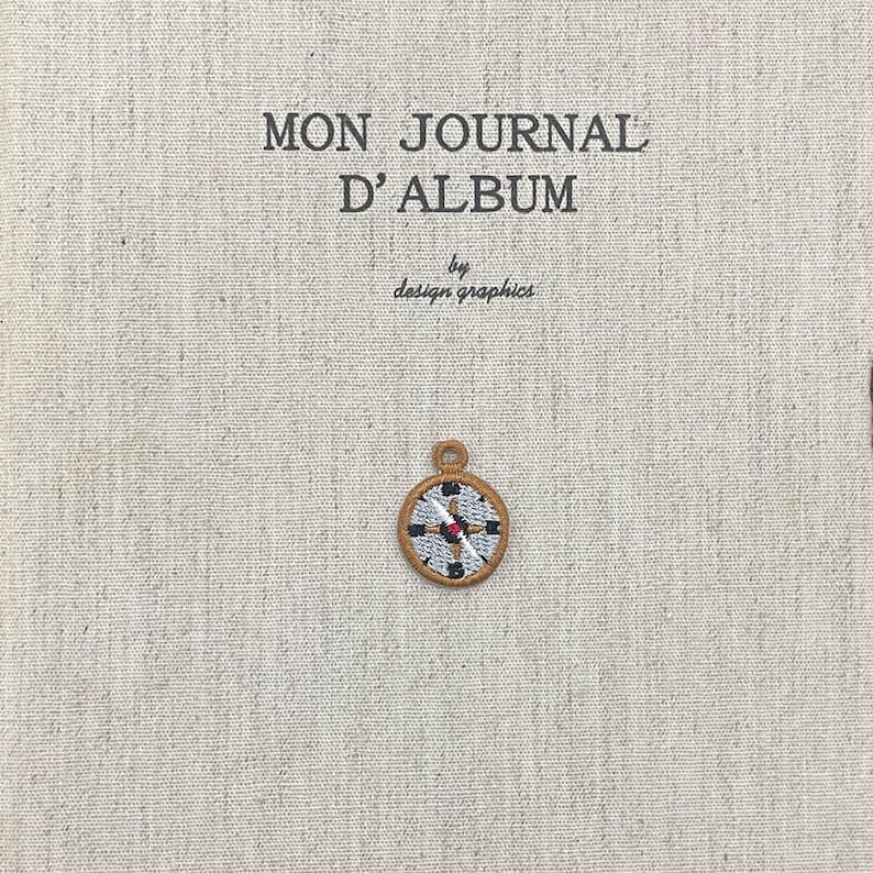 May include: A beige fabric with the text "MON JOURNAL D'ALBUM" and "by design graphics" printed in black. A small, embroidered compass charm with red, black, and white details is attached to the fabric.