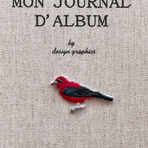 May include: Embroidered patch featuring a vibrant red bird with black wings and tail, perched on a textured beige fabric. The text "MON JOURNAL D'ALBUM" and "by design graphics" are above the bird.