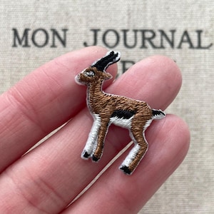 May include: Embroidered patch of a gazelle in brown, black, and white. The gazelle is standing with its legs slightly apart. The patch is detailed with intricate stitching, and the background has the text "MON JOURNAL".