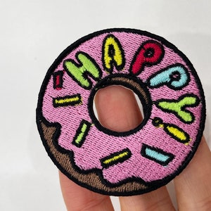Op de afbeelding: Geborduurde patch in de vorm van een roze donut met kleurrijke hagelslag. Het woord "HAPPY" is in verschillende kleuren over het midden van de donut geborduurd. De patch heeft een zwarte rand.