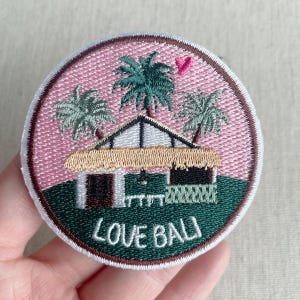 May include: Embroidered patch depicting a tropical scene with a hut, palm trees, and the text "LOVE BALI." The patch has a pink background with a green base and a brown border. A small pink heart is above the hut.