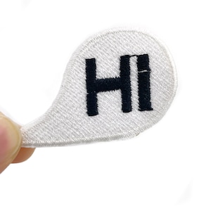 Pode incluir: Patch bordado branco em forma de balão de fala com a palavra "HI" em letras pretas. O patch é segurado por uma mão contra um fundo branco. O patch é provavelmente para roupas ou acessórios.