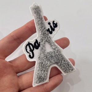 May include: Embroidered patch of the Eiffel Tower in grey, with the word "Paris" in black script. The patch has a white border and a textured, fuzzy appearance. Suitable for decorating clothing or accessories.