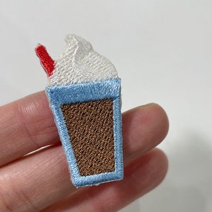 May include: Embroidered patch of a milkshake with a red straw. The milkshake has a white whipped cream topping, a light blue rim, and a brown cup. The patch is a fun accessory for clothing or bags.