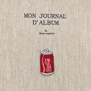 以下が含まれることがあります： ベージュ色の生地のジャーナルカバー。黒で「MON JOURNAL D'ALBUM」と「by design graphics」の文字が書かれています。赤いコカ・コーラの缶のパッチがカバーに縫い付けられています。