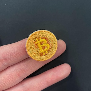 May include: Embroidered gold Bitcoin patch with a white border. The Bitcoin symbol is in the center of the patch. The patch is round and has a textured appearance.