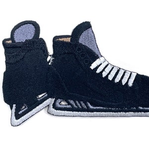 Könnte beinhalten: Gestickter Aufnäher mit zwei schwarzen Eislaufschuhen mit weißen Schnürsenkeln und silbernen Kufen. Die Schlittschuhe haben eine strukturierte Optik und sind vor weißem Hintergrund platziert. Für Eishockeyfans.