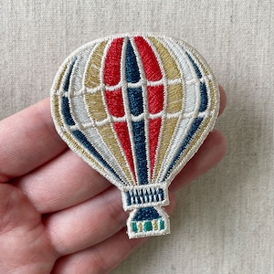 以下が含まれることがあります： カラフルなデザインの刺繍入り熱気球パッチ。バルーンは赤、金、白、ネイビーブルーのセクションが特徴です。パッチは、衣類やアクセサリーに追加するのに適した、詳細なステッチの外観をしています。