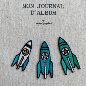 May include: Three embroidered rocket patches in blue, white, and green, each with a star emblem. The patches are on a textured, beige fabric. Text at the top reads "MON JOURNAL D'ALBUM" and "by design graphics".