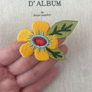 以下が含まれることがあります： 赤と青の中心と緑の葉が付いた、刺繍された黄色の花のパッチ。パッチは、背景に「D'ALBUM by design graphics」というテキストが入った花のデザインです。