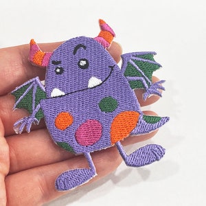 Puede incluir: Parche bordado con un monstruo morado con cuernos naranjas, alas verdes y lunares coloridos. El monstruo tiene una expresión sonriente y dientes blancos. Este parche caprichoso es adecuado para ropa o accesorios.