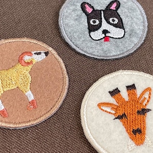 Può includere: Tre toppe rotonde in feltro ricamato con disegni di animali: un cane, un bulldog francese e una giraffa. Le toppe hanno un bordo cucito e sono poste su uno sfondo marrone.