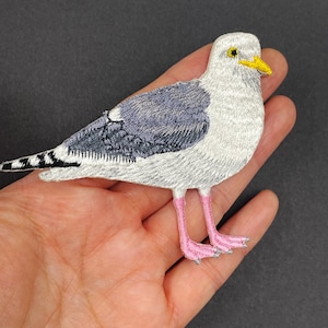 May include: An embroidered patch shaped like a seagull. The bird features white plumage on the head and chest, dark gray wings with black-tipped feathers, a yellow beak with a small black eye, and bright pink legs with webbed feet.
