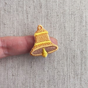 Puede incluir: Parche de campana bordada en tonos dorados y marrones. La campana tiene un diseño a rayas y un pequeño lazo en la parte superior. El parche es probablemente para fines decorativos, como en ropa o accesorios.