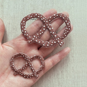 Op de afbeelding: Twee bruine pretzel patches met witte stippen. De patches zijn geborduurd met een gedetailleerd ontwerp. Eén patch is groter dan de andere. De patches zijn een ambachtelijk item met een voedselthema.