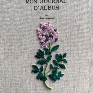 Op de afbeelding: Geborduurde bloemenpatch met roze en paarse bloemen, groene bladeren en stengel, op een beige stof. De tekst bovenaan luidt "MON JOURNAL D'ALBUM" en "design graphics" eronder. Perfect voor handwerk en decoratie.