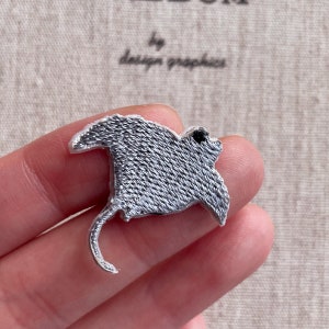 May include: Embroidered patch featuring a detailed stingray design. The patch has a textured, gray body with a silver outline. The design includes the text "by design graphics" in a script font.