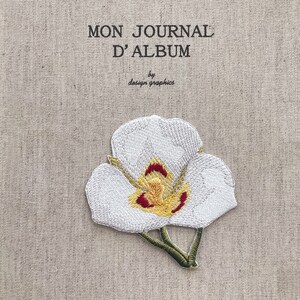 May include: A beige fabric journal cover with the text "MON JOURNAL D'ALBUM" and "by design graphics". An embroidered white orchid with yellow and red accents is featured on the cover.