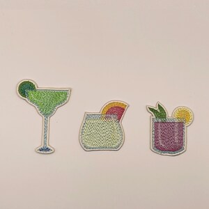May include: Three embroidered patches featuring cocktail designs. The first is a green margarita with a lime slice. The second is a light green drink with an orange slice. The third is a purple drink garnished with green leaves and a yellow lemon slice.
