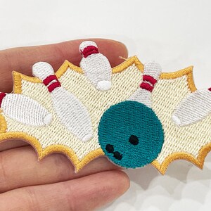 May include: Embroidered patch featuring a bowling scene. The design shows a teal bowling ball hitting white bowling pins with red stripes, set against a yellow starburst background. A fun accessory for clothing or bags.