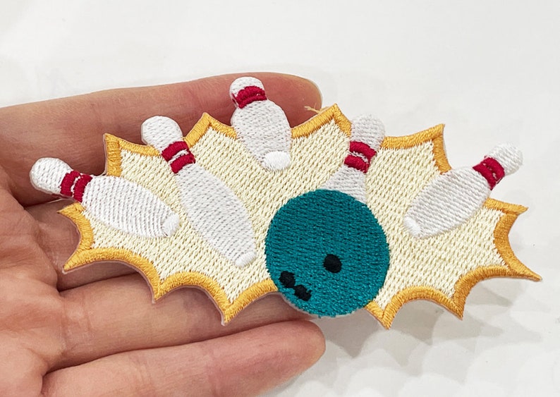 May include: Embroidered patch featuring a bowling scene. The design shows a teal bowling ball hitting white bowling pins with red stripes, set against a yellow starburst background. A fun accessory for clothing or bags.
