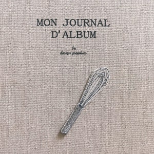 May include: A beige fabric journal cover with the text "MON JOURNAL D'ALBUM" and "by design graphics" in black. A silver whisk embroidery is on the cover.