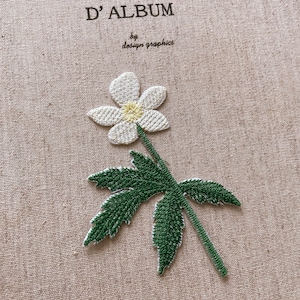 May include: Embroidered flower applique with a white flower and green leaves on a tan fabric background. The flower has a yellow center. The text "D'ALBUM by design graphics" is at the top.