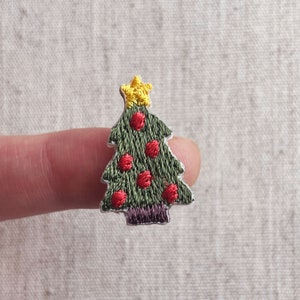 Może przedstawiać: Haftowana naszywka z choinką. Naszywka przedstawia zielone drzewko z czerwonymi bombkami i żółtą gwiazdą na górze. Drzewko ma fioletową podstawę i srebrny kontur.