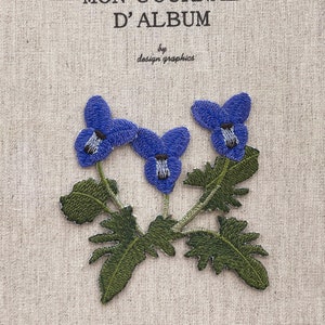 May include: Embroidered blue flower patch with green leaves and stems. The patch features three blue flowers with detailed stitching and a textured appearance. The background is a light beige fabric. Text at the top reads "MON JOURNAL D'ALBUM" and "by design graphics".