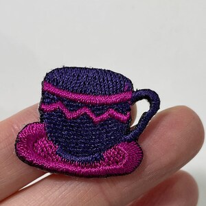 May include: Embroidered patch of a teacup and saucer. The teacup is dark blue with a pink zig-zag design and a pink stripe. The saucer is pink. The patch is small and detailed.