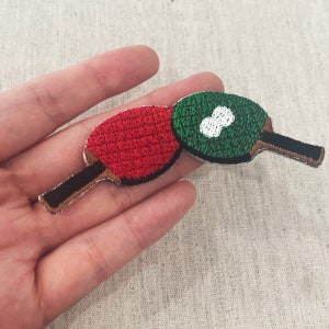 Könnte beinhalten: Gestickter Aufnäher mit zwei Tischtennisschlägern. Einer ist rot mit schwarzem Griff, der andere grün mit einem weißen Tischtennisball-Design und einem schwarzen Griff. Die Aufnäher befinden sich auf einem neutralen Stoffhintergrund.