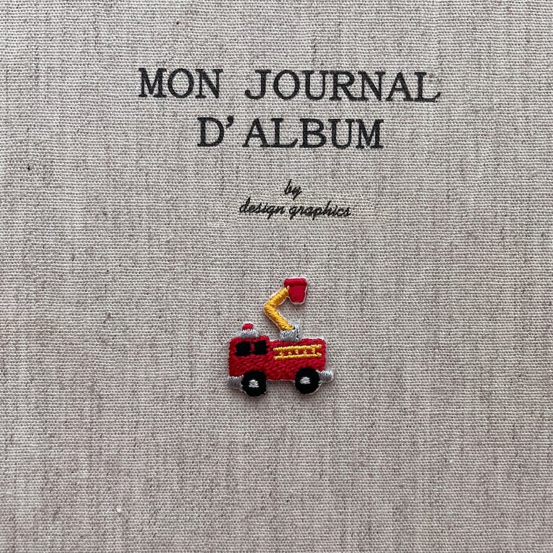 May include: Embroidered journal cover with the text "MON JOURNAL D'ALBUM" and "by design graphics". A small, red fire truck with yellow details is embroidered on the cover. The background is a textured, light beige fabric.