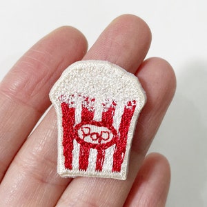 May include: Embroidered patch in the shape of a popcorn container. The patch features a white popcorn design with red stripes and the word "POP" in a red oval.