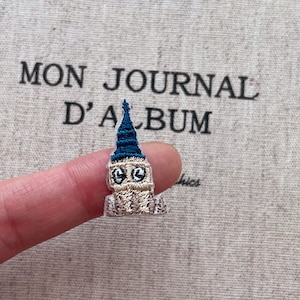 Op de afbeelding: Geborduurde Big Ben-patch met een blauwe bovenkant en beige lichaam. De patch wordt door een vinger vastgehouden voor een achtergrond met de tekst "MON JOURNAL D'ALBUM".