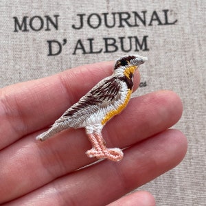 Pode incluir: Remendo de pássaro bordado com detalhes em fio marrom, branco, amarelo e pêssego. O remendo apresenta um design detalhado de um pássaro com as asas dobradas. O fundo inclui o texto "MON JOURNAL D'ALBUM".