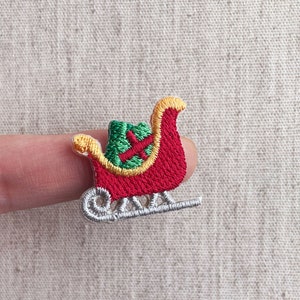 Puede incluir: Parche bordado de trineo navideño. El trineo es rojo con ribetes dorados y un asiento verde. Los patines son plateados. Una decoración festiva para ropa, bolsos o manualidades.
