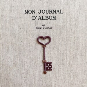 Op de afbeelding: Geborduurde sleutel in bordeauxrood, met een hartvormige bovenkant en gedetailleerde versieringen. De sleutel is afgebeeld op een beige stoffen achtergrond. De Franse tekst "MON JOURNAL D'ALBUM" en "by design graphics" staan erboven.
