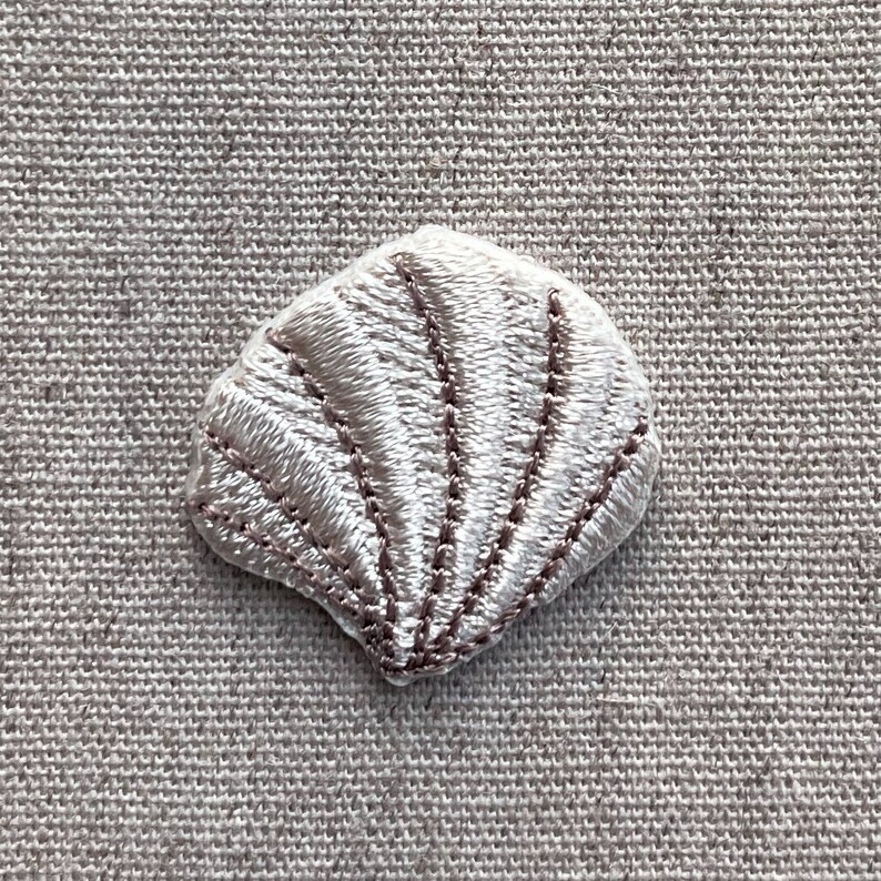 K&ouml;nnte beinhalten: Ein kleiner, gestickter Muschel-Aufn&auml;her in Wei&szlig;- und Hellbraunt&ouml;nen. Der Aufn&auml;her weist detaillierte Stiche auf, die die Form und die Rippen der Muschel umrei&szlig;en. Der Aufn&auml;her dient wahrscheinlich dekorativen Zwecken.