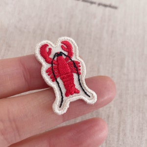 Op de afbeelding: Geborduurde patch met een rood kreeftontwerp en zwarte details. De kreeft staat tegen een crèmekleurige achtergrond met een gestikte rand. Geschikt voor kleding en accessoires.