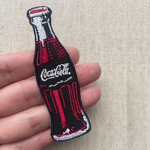 以下が含まれることがあります： コカ・コーラのボトルの形をした刺繍パッチ。パッチは、白いスクリプトのコカ・コーラロゴを含む、黒い背景に赤と白のアクセントが特徴です。ボトルの形は白で縁取られています。