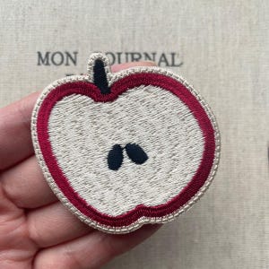 May include: Embroidered apple patch with a cream interior, a dark red outline, and black seeds. The patch is in the shape of a half apple. The text "MON JOURNAL" is visible in the background.