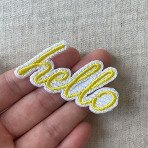 Peut inclure: Un écusson brodé blanc avec le mot "hello" en écriture jaune. Le patch a une bordure jaune et se trouve sur un fond de tissu de couleur neutre.