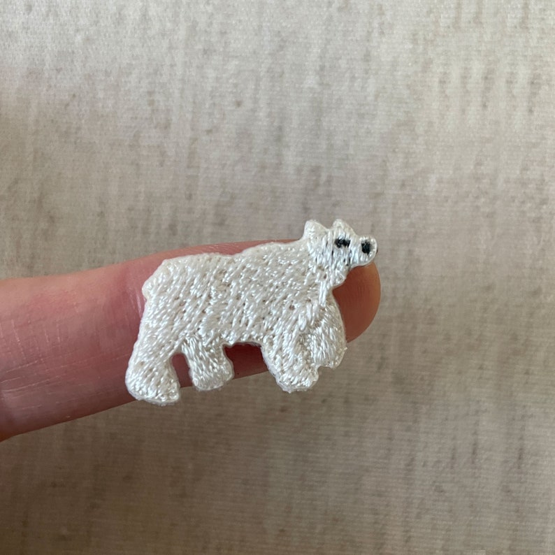 K&ouml;nnte beinhalten: Gestickter wei&szlig;er Eisb&auml;r-Aufn&auml;her mit schwarzen Augen und Nase. Der Aufn&auml;her besteht aus einem strukturierten, gewebten Material. Der B&auml;r wird in stehender Position gezeigt.