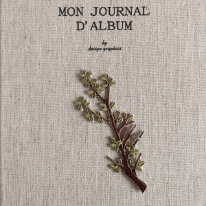 Op de afbeelding: Een beige tijdschriftomslag met de Franse tekst "MON JOURNAL D'ALBUM" en "by design graphics". Een bruine geborduurde tak met groene bladeren versiert de omslag.