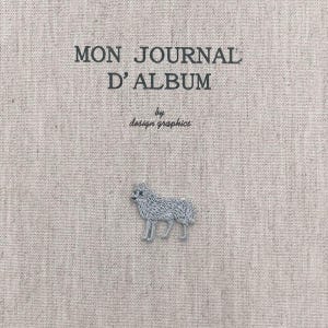May include: A beige journal cover with the text "MON JOURNAL D'ALBUM" and "by design graphics" in black. A small, embroidered grey dog is at the bottom. The cover has a textured appearance.