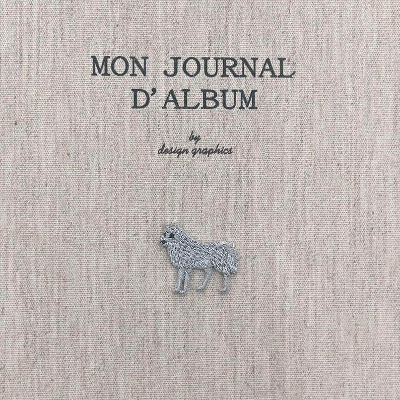May include: A beige journal cover with the text "MON JOURNAL D'ALBUM" and "by design graphics" in black. A small, embroidered grey dog is at the bottom. The cover has a textured appearance.