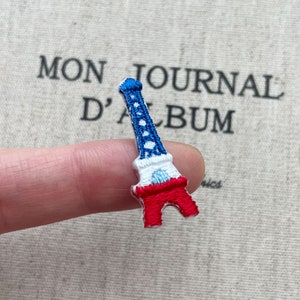 May include: Embroidered Eiffel Tower patch in red, white, and blue. The patch is held by a finger, with the text "MON JOURNAL D'ALBUM" in the background. The patch is a miniature representation of the iconic Parisian landmark.