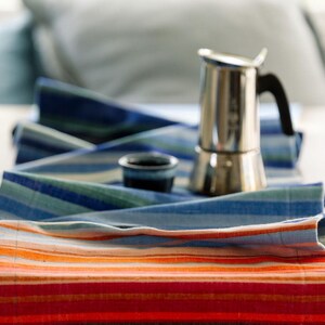 Puede incluir: Un camino de mesa largo y rectangular con rayas horizontales en tonos vibrantes de naranja, rojo, azul y verde. Sobre el camino de tela se encuentran una cafetera italiana de metal plateado con asa negra y una pequeña taza de cerámica azul.